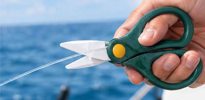 Green-handled ceramic fishing scissors cutting fishing line against a saltwater fishing background.